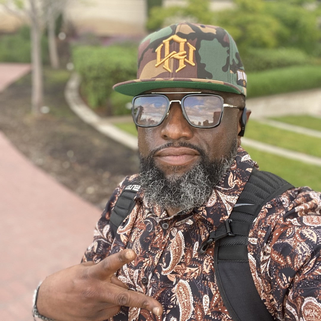Joyful man posing outdoors, wearing a RENOWNED WEAR Camo Snapback hat exuding positivity.